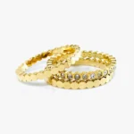 Cette image représente sur fond blanc trois anneaux positionné sur leurs tranches. Ce bijou pour femme est de couleurs or avec des rebords ciseler en forme d'ourlets régulier, comme un design en vagues. L'anneau bague du milieu est sur son rebord incrusté de façon régulière de petit zirconium blanc.